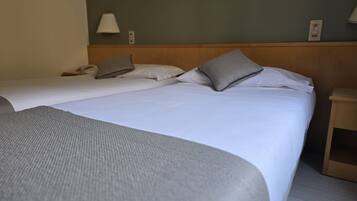 Een tweepersoonsbed met witte lakens en twee kussens, een nachtkastje en een lamp aan de muur.