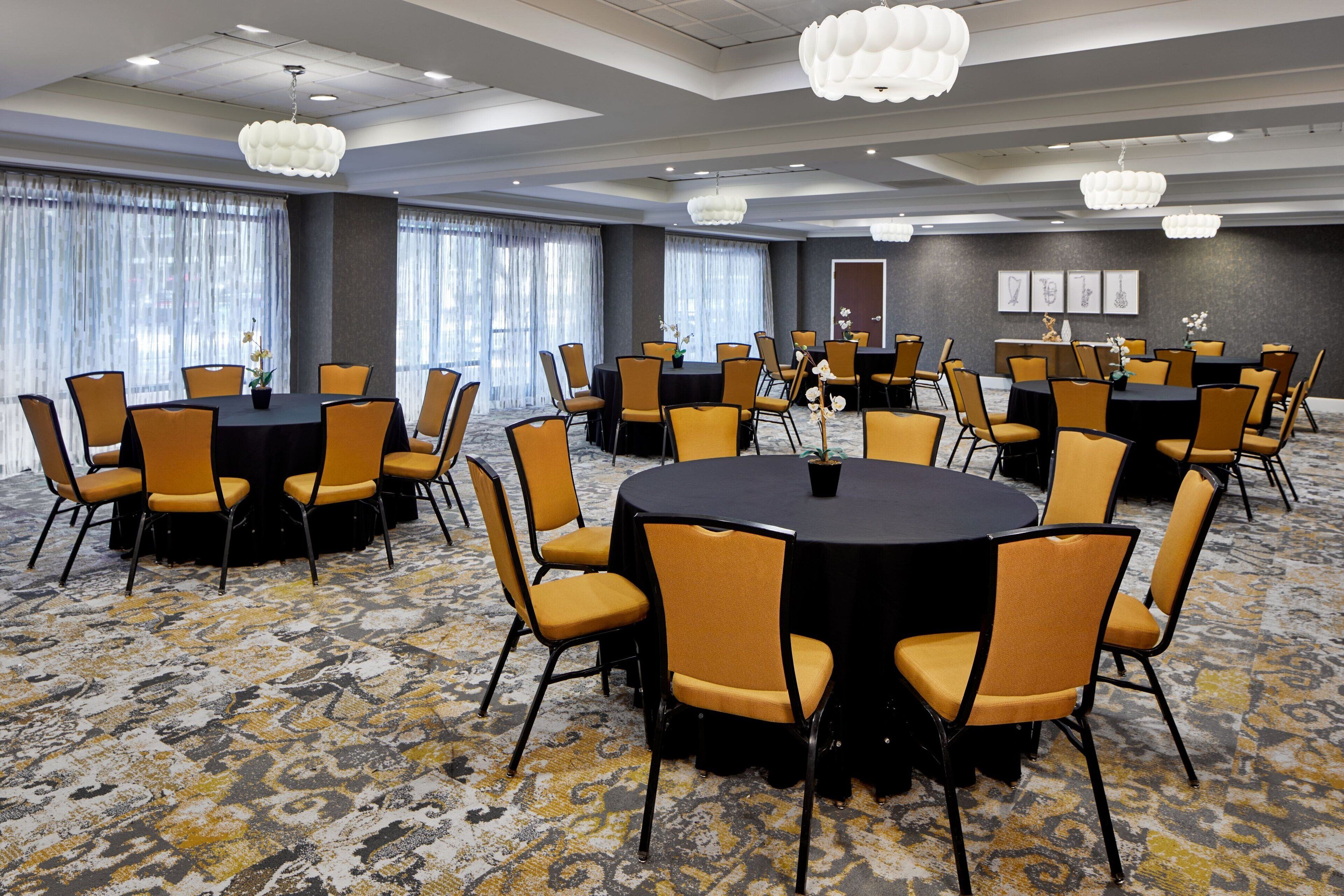 Un salón de conferencias con varias mesas redondas y sillas, ventanas amplias con cortinas y una alfombra estampada.
