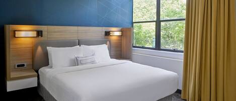 Habitación de hotel con una cama grande, cabecera con iluminación integrada, mesita de noche y ventana con cortinas.