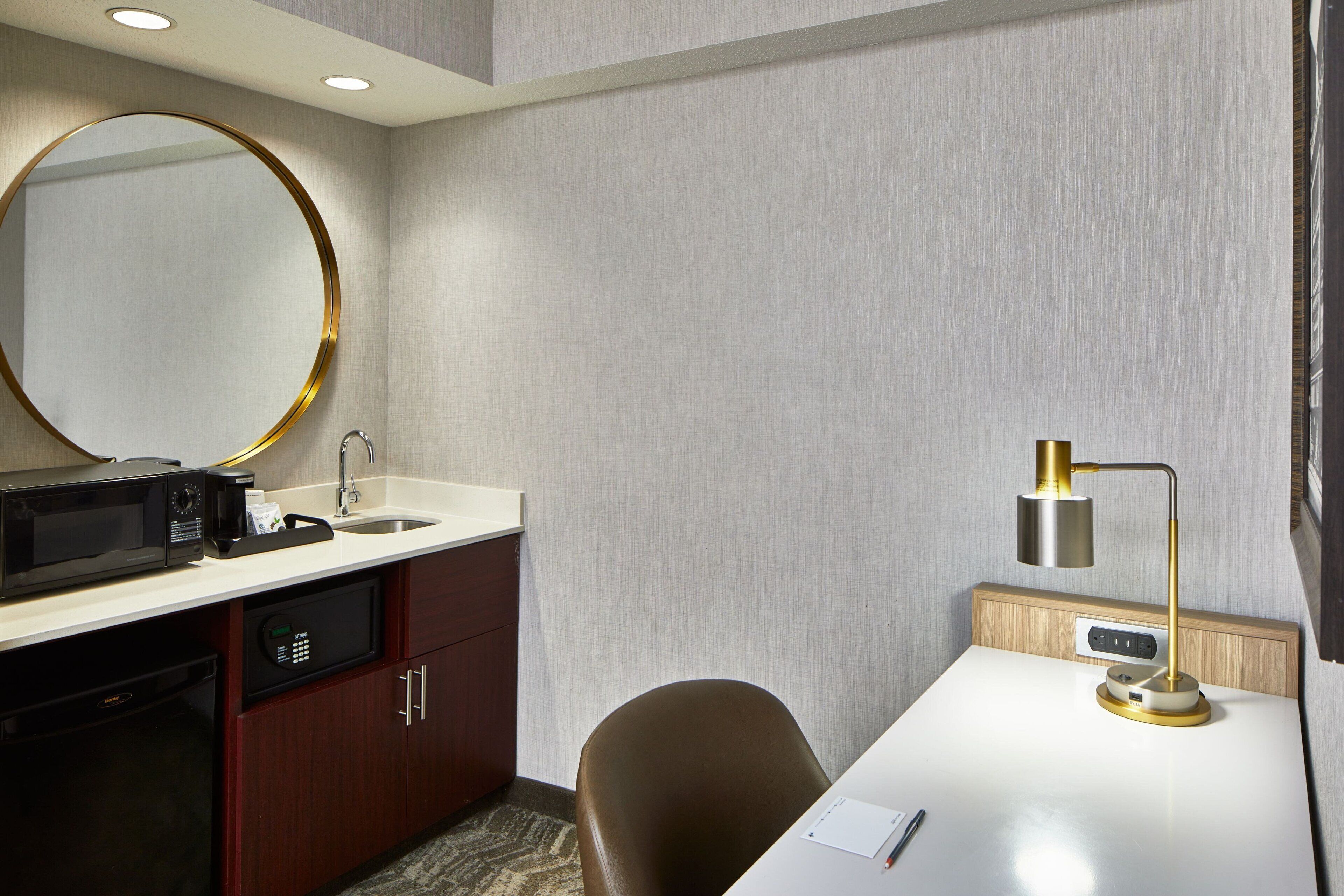 A hotel room with a white desk, a brown chair, a round mirror, a sink, a microwave, a coffee maker, and a bedside lamp.