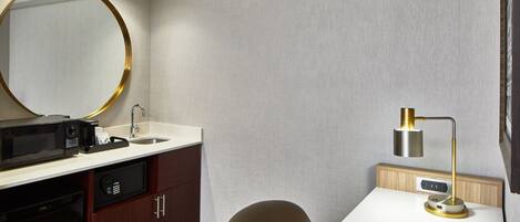 A hotel room with a white desk, a brown chair, a round mirror, a sink, a microwave, a coffee maker, and a bedside lamp.