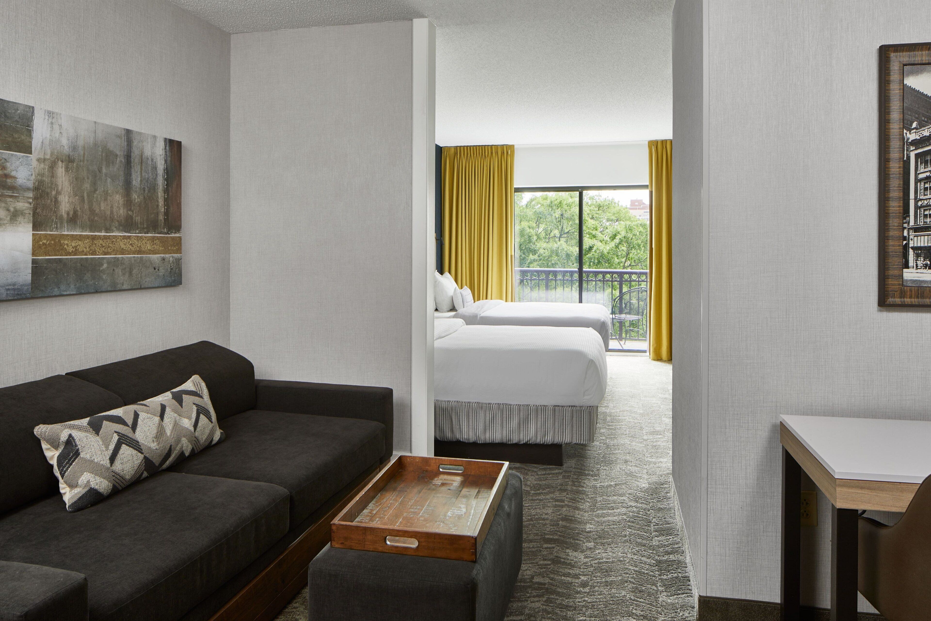 Habitación de hotel con sofá, una mesita de madera, cama con dos almohadas y vista a un balcón con cortinas amarillas.