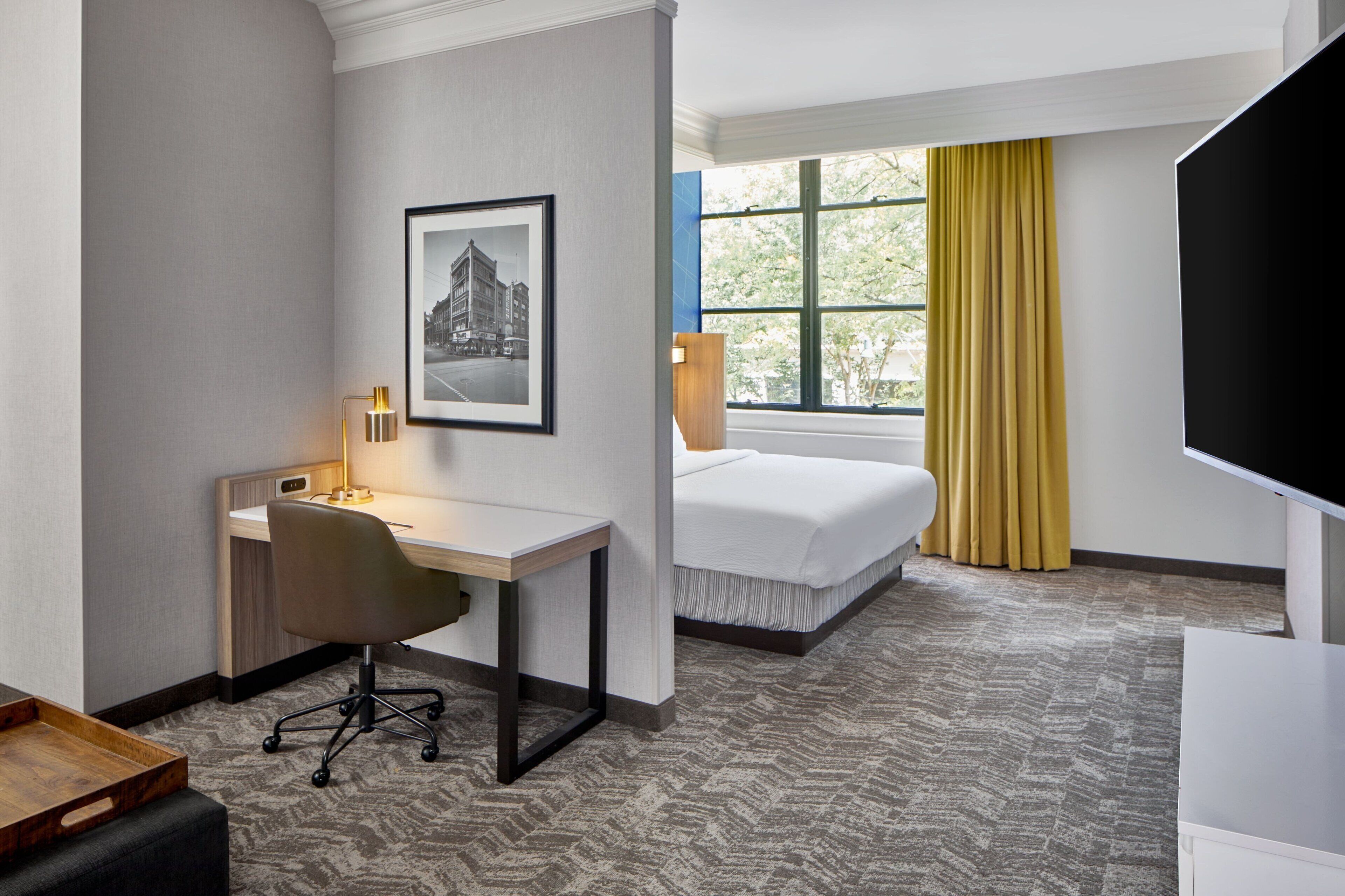 Habitación de hotel con escritorio, silla, cama y un ventanal con cortinas amarillas.