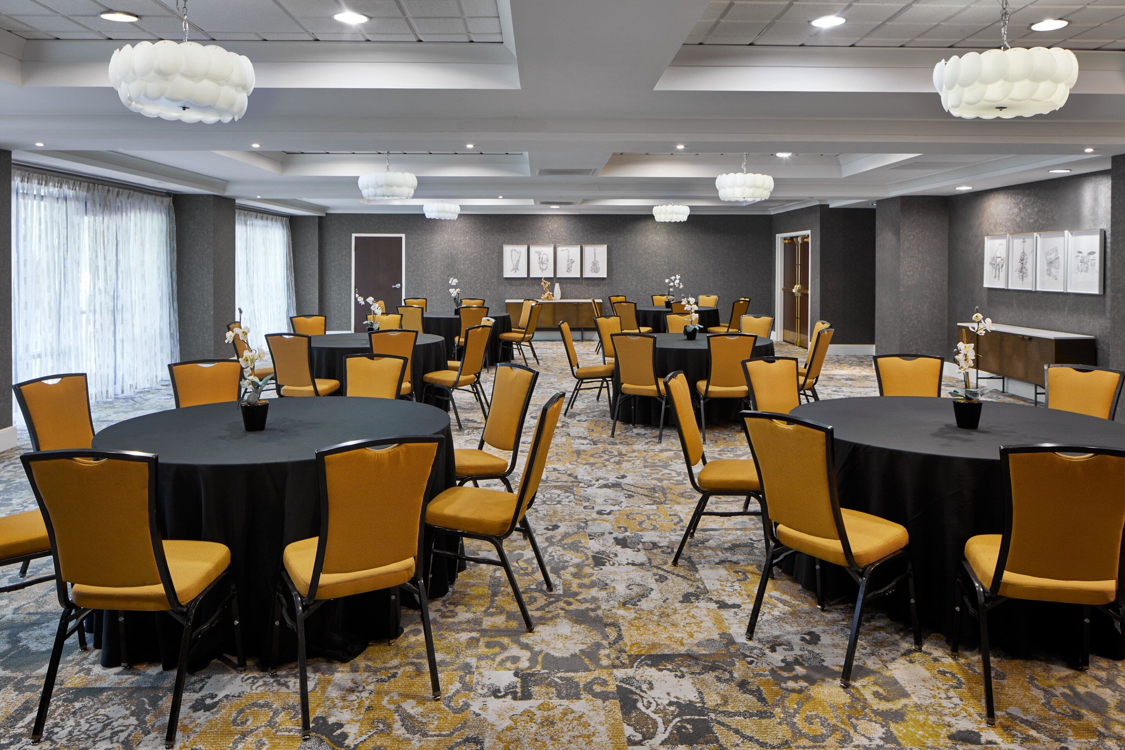A conference room with multiple round tables and chairs, large windows with curtains, and a patterned carpet.