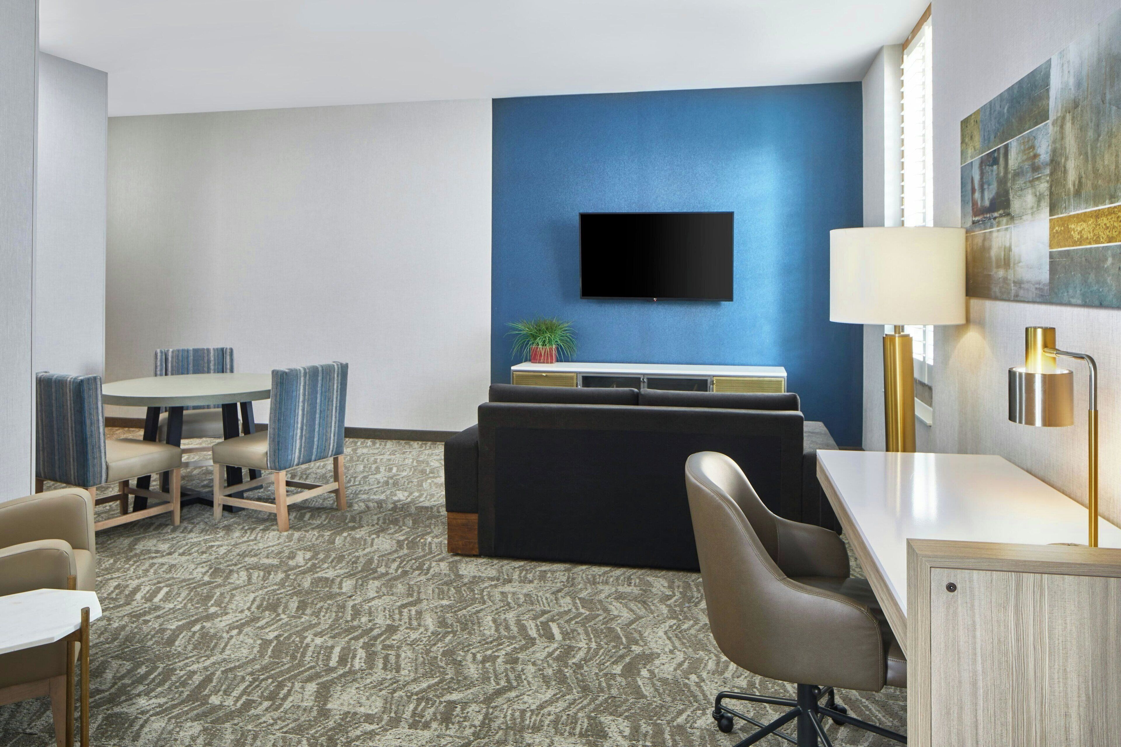 A hotel room with a flat-screen TV, a sofa, a dining table with chairs, a lamp, and a carpeted floor.