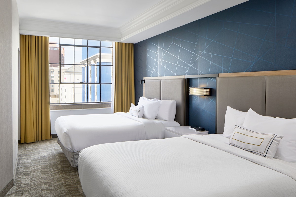 A hotel room with two beds, a large window with curtains, a geometric wall design, and a nightstand with a lamp.