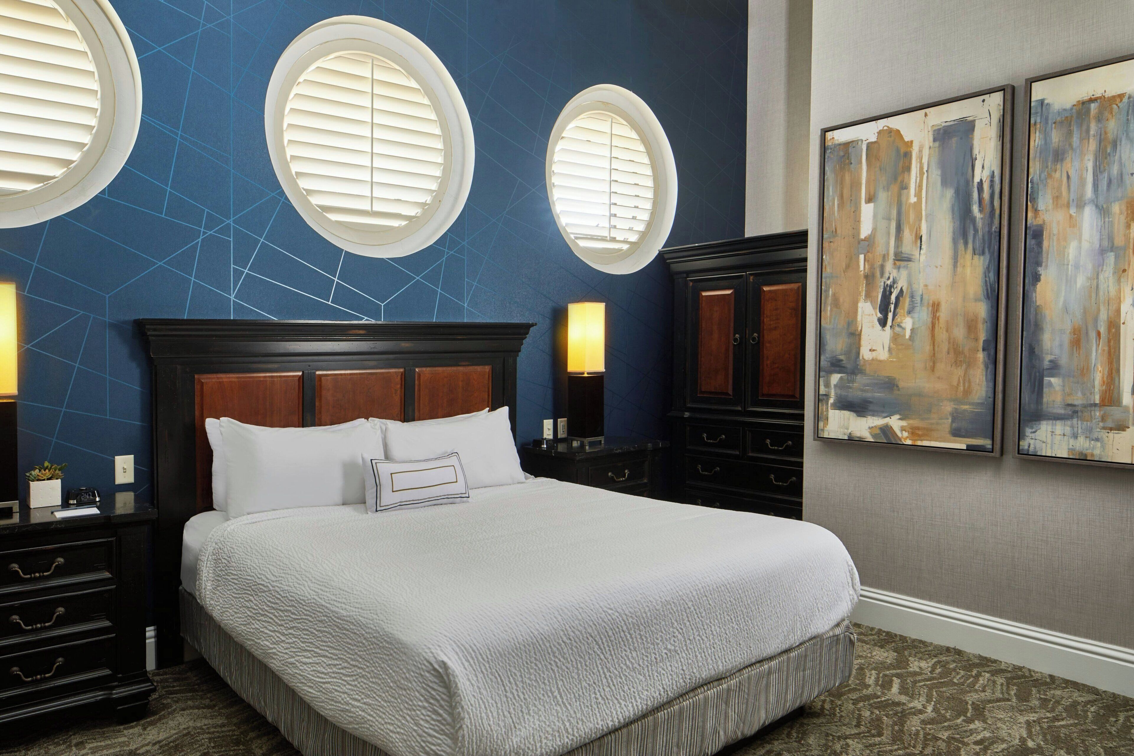Une chambre d’hôtel avec un grand lit, deux tables de chevet avec des lampes, une commode et trois fenêtres rondes fixées au mur.