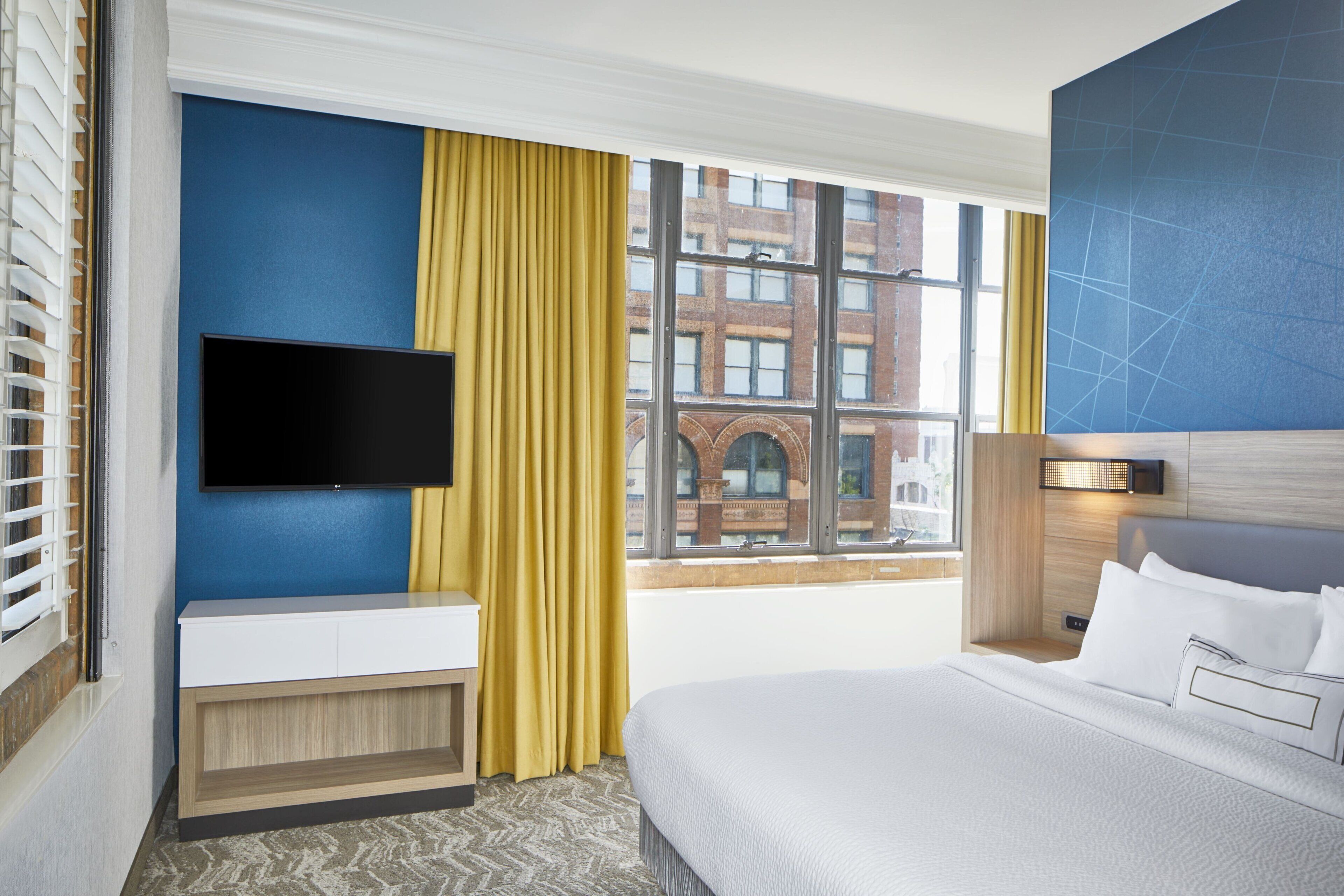 A hotel room with a bed, a bedside table, a TV mounted on the wall, and a window with curtains.