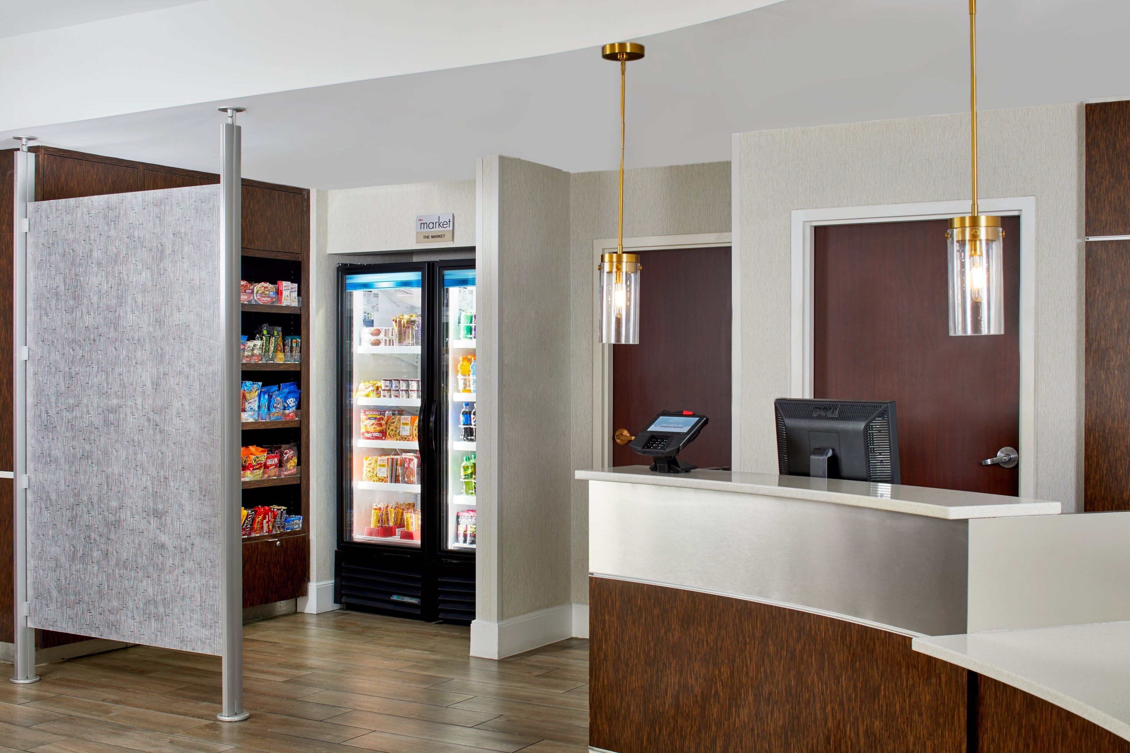A hotel reception area with a self-service kiosk, a refrigerator stocked with snacks, and a wooden partition.