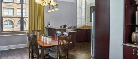 A dining area with a wooden table and chairs, a kitchen in the background, and a large window with curtains.