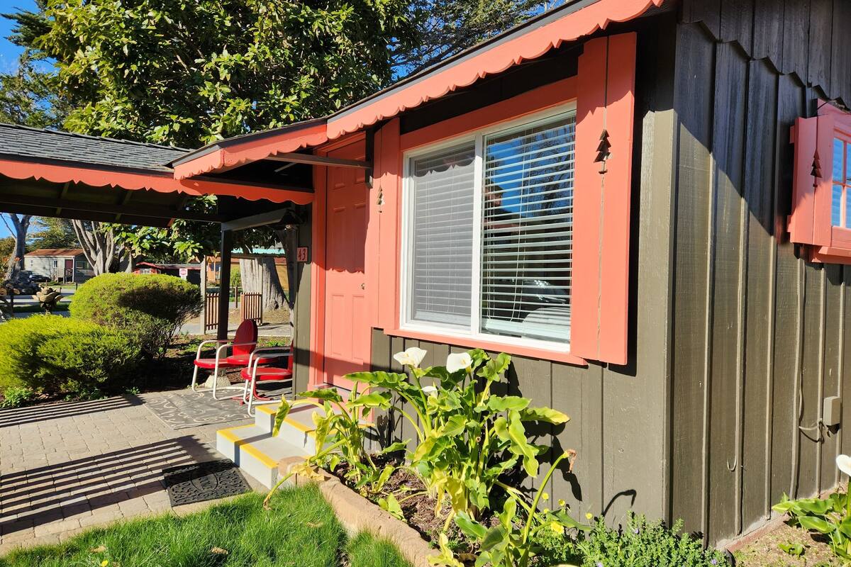 Ein Haus mit roter Fassade, ein Fenster mit Jalousien, ein roter Stuhl draußen und ein kleiner Garten mit Pflanzen und einem Weg.