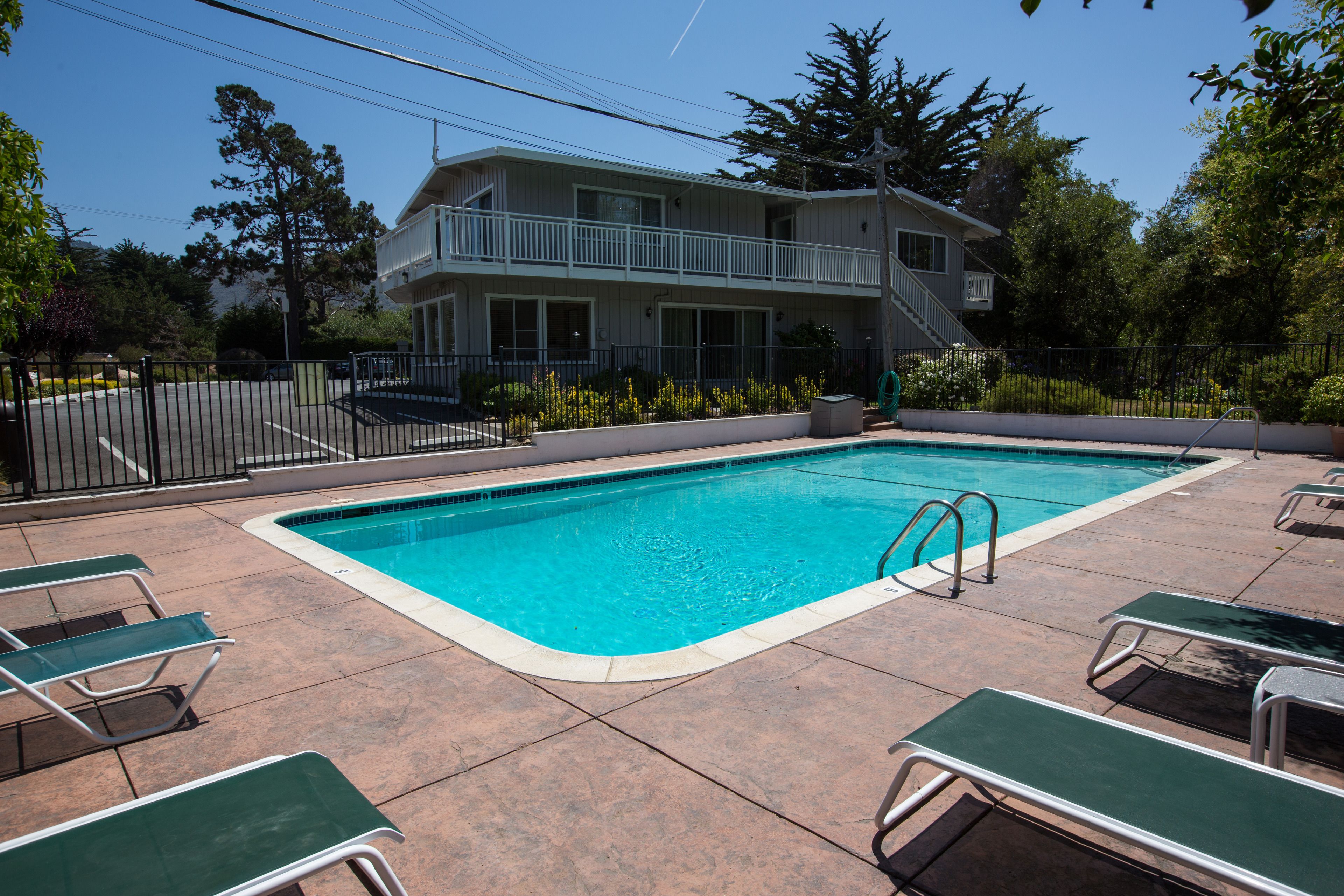 Une piscine avec des chaises longues, un bâtiment à deux étages doté d’un balcon, et une végétation luxuriante tout autour.
