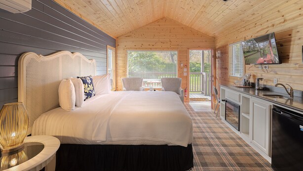 A wooden cabin bedroom with a bed, bedside table, and a view of the outside. - Carmel River Inn & Cottages (Carmel)