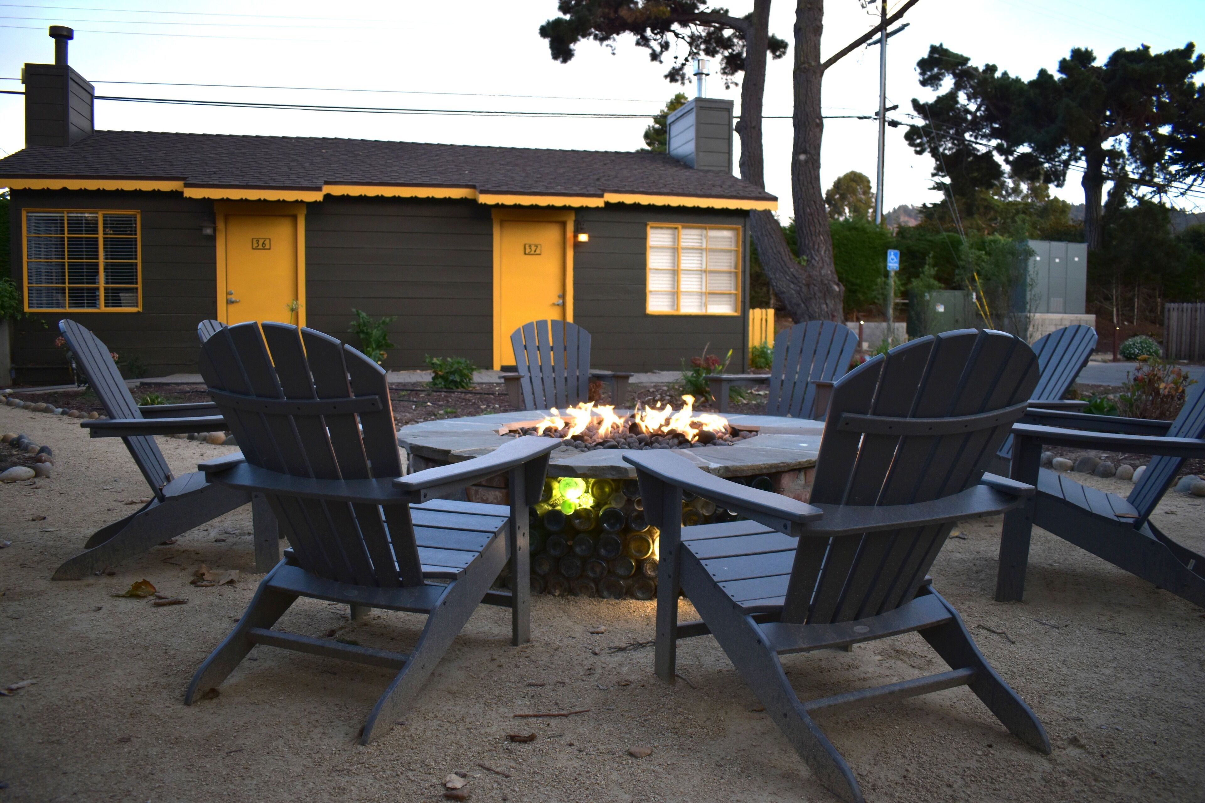 A cozy outdoor seating area with six Adirondack chairs around a central fire pit, set against a house with a yellow door and a number 32 sign.