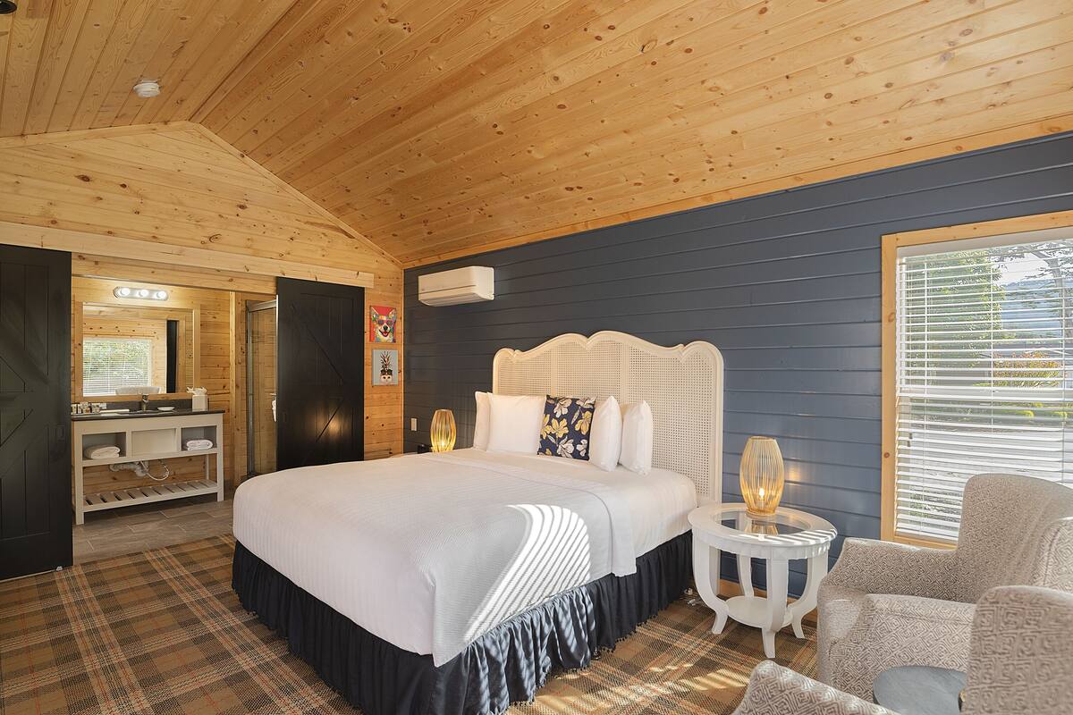 Ein Schlafzimmer mit einem großen Bett, einem Nachttisch, einem Sessel, einem Schminktisch mit Spiegel und einem Fenster mit Jalousien.