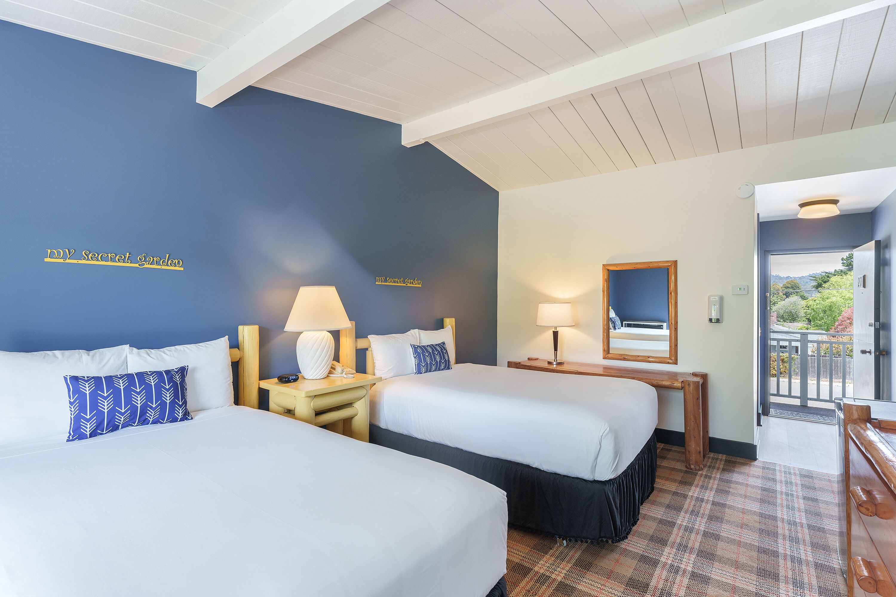 A hotel room with two beds, a wooden bench, a desk, and a view of the outdoors.