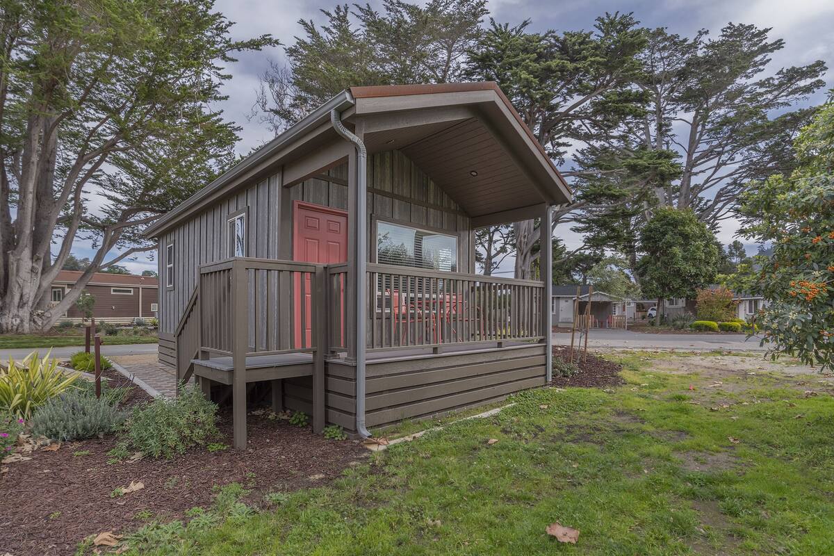 Eine kleine Holzhütte mit roter Tür, einer Veranda und Blick auf eine Wohngegend mit Bäumen und Häusern.