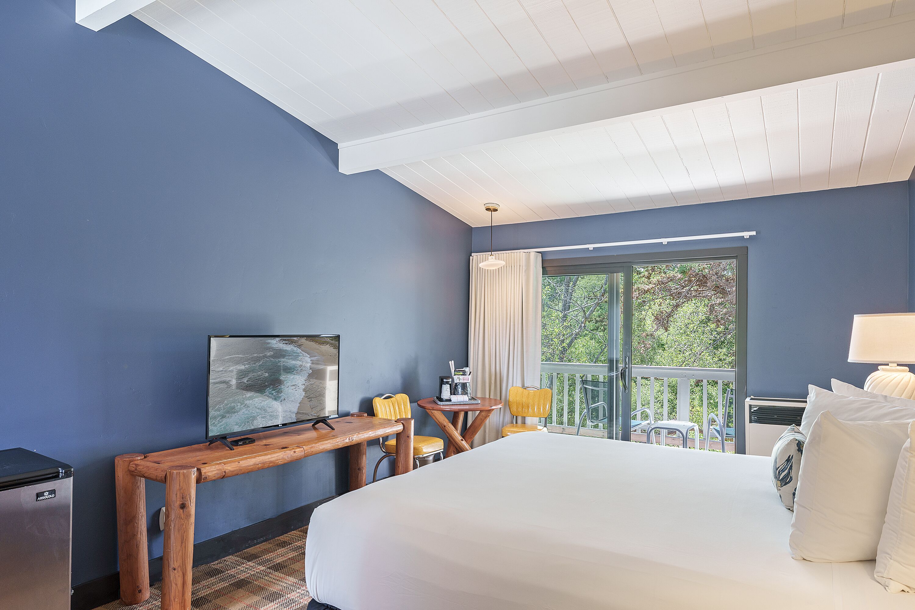 Quarto de hotel com uma cama grande, uma escrivaninha de madeira com televisão, uma varanda com cadeiras e uma janela com cortinas.