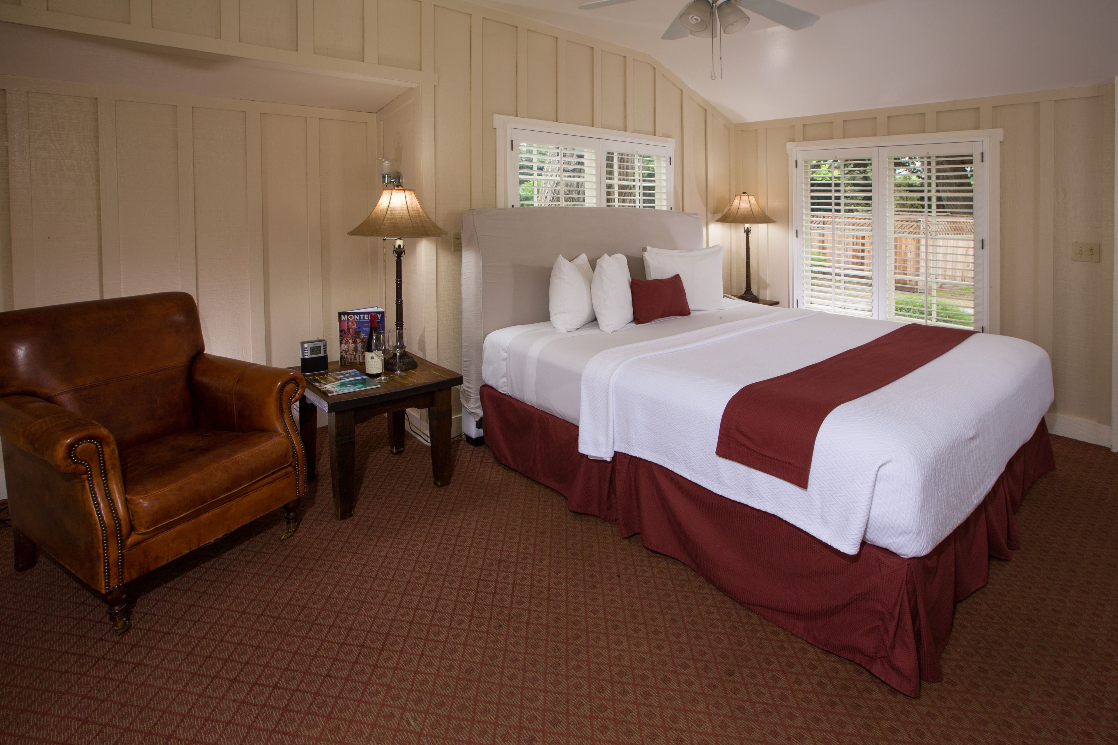 A hotel room with a large bed, a leather armchair, a bedside table with a lamp, and a window with shutters.