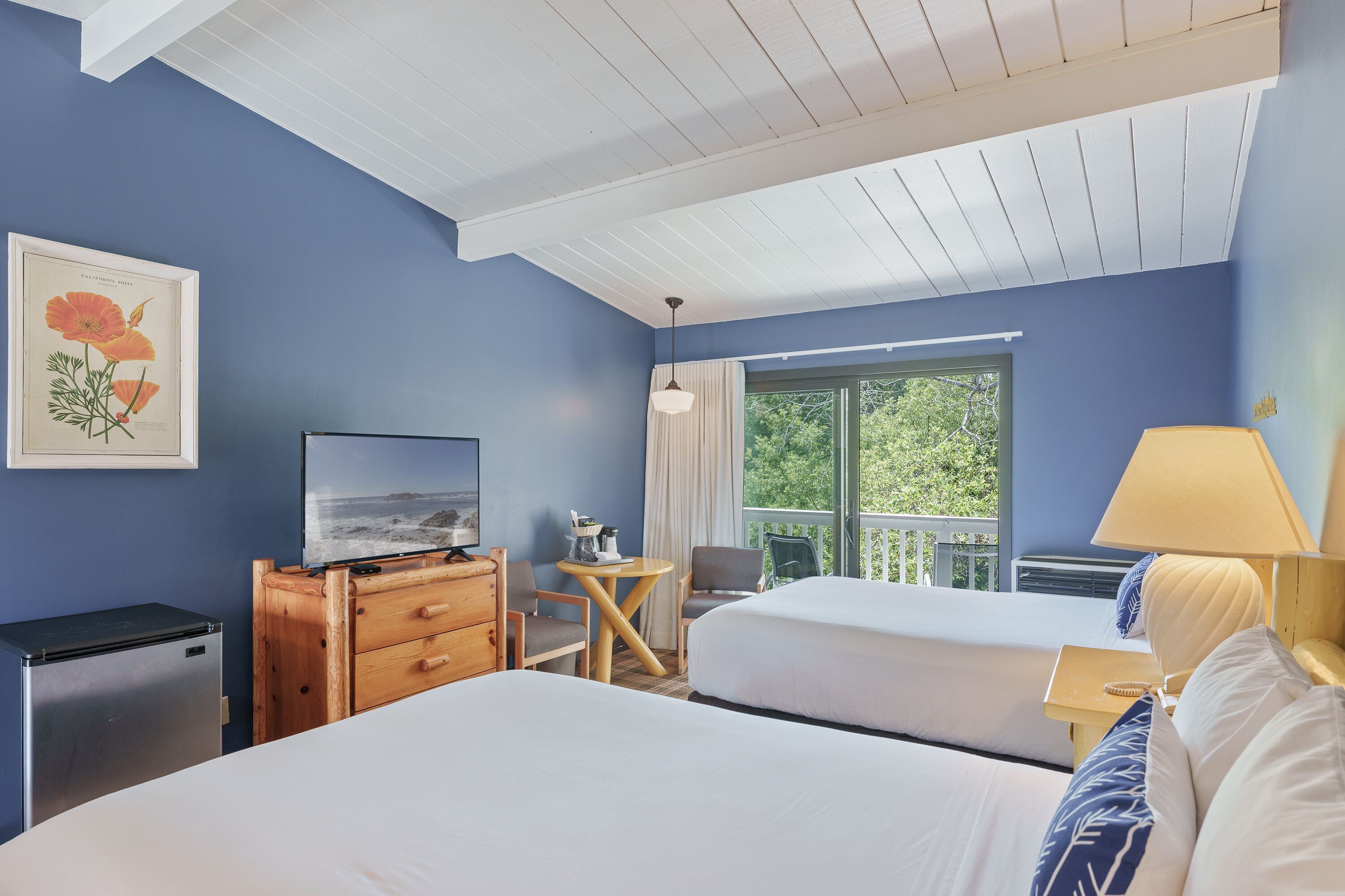 A hotel room with two beds, a TV, a wooden dresser, a small table, and a balcony with a view of greenery.