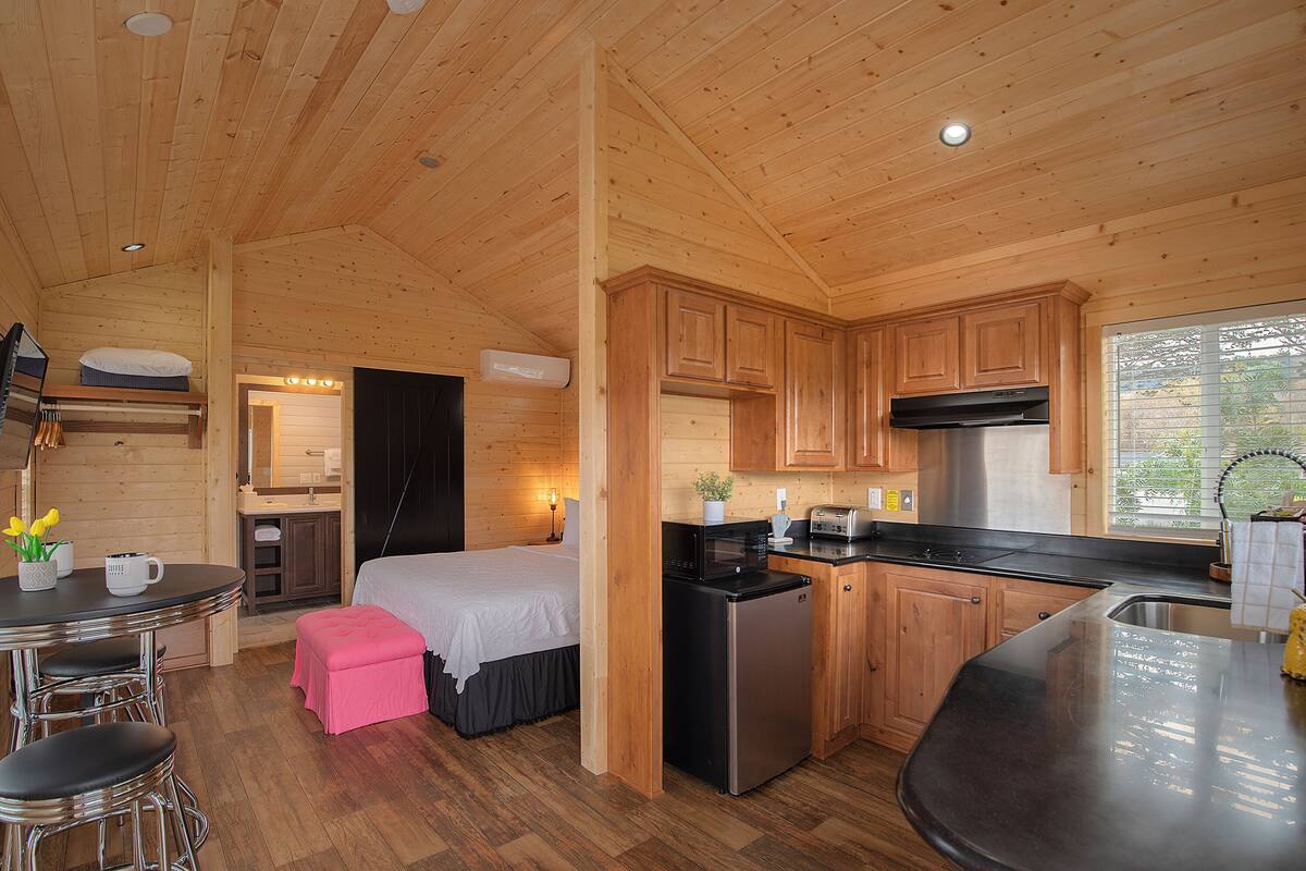 Eine kompakte Holzhütte mit einer Küchenzeile, einem kleinen Essbereich und einem Schlafzimmer mit einem Bett und einem pinken Pouf.