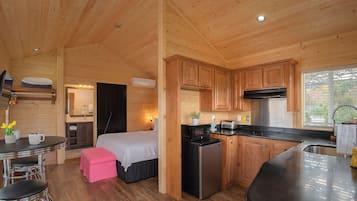 A compact wooden cabin with a kitchenette, a small dining area, and a bedroom with a bed and a pink ottoman.