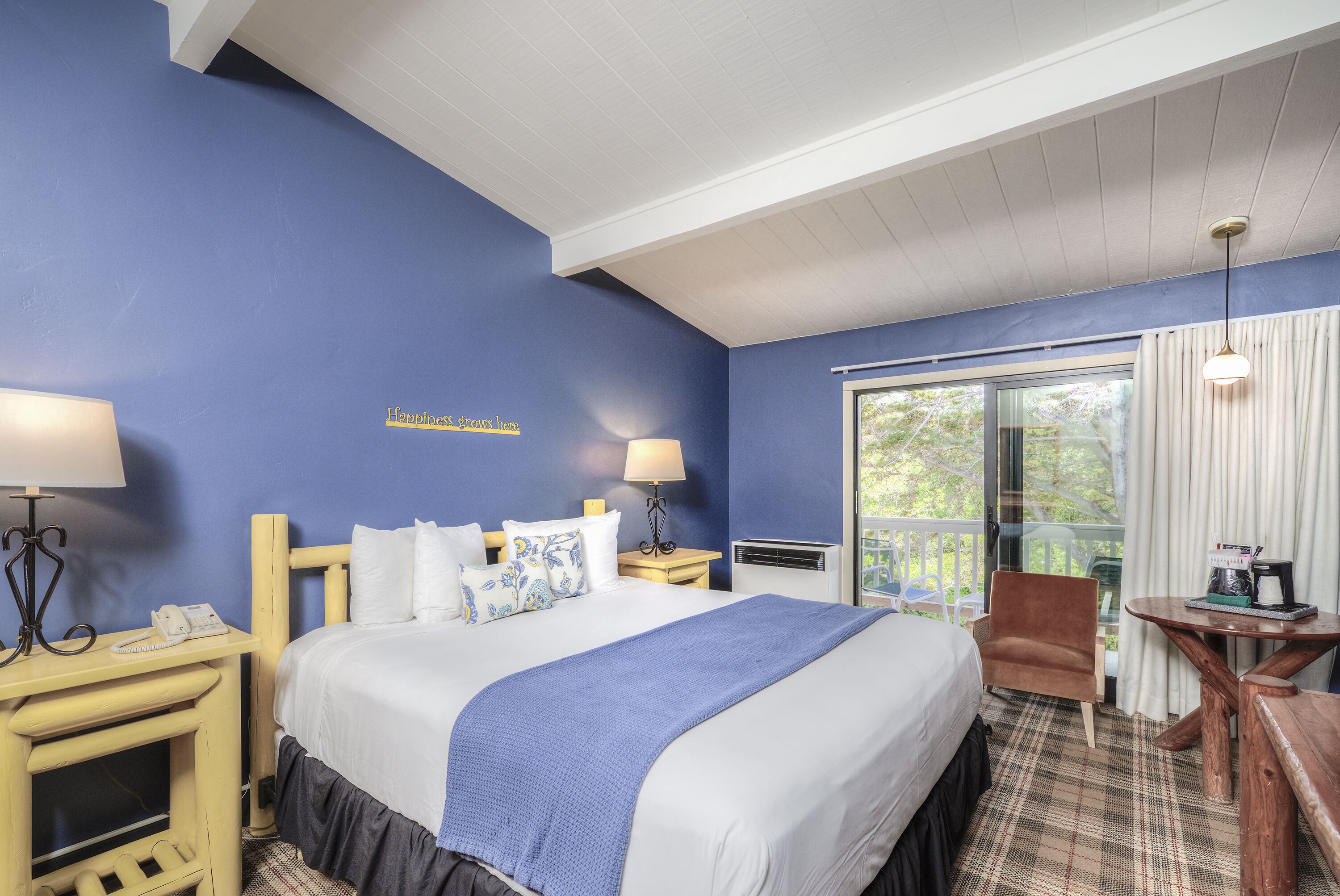 A hotel room with a large bed, two bedside tables with lamps, a wooden chair, a small table with a coffee maker, and a view of greenery through the window.
