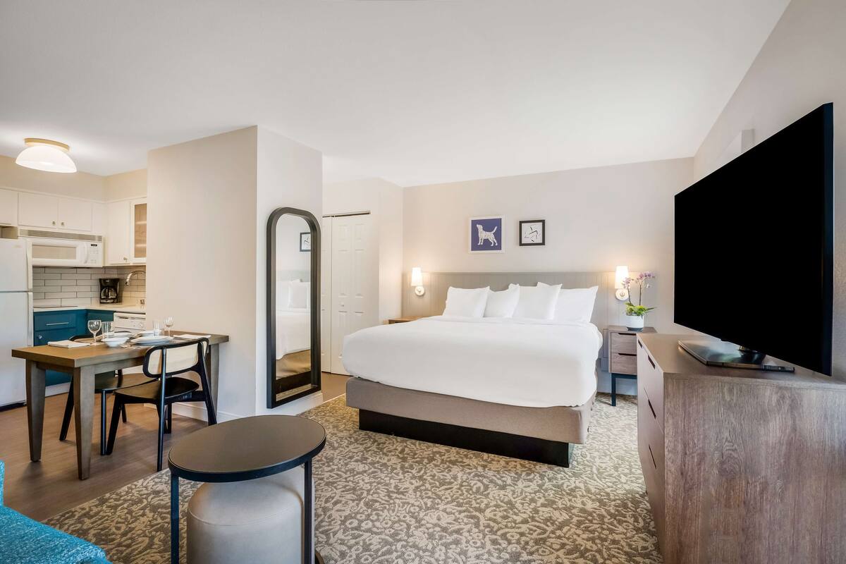 Habitación de hotel con cama, escritorio, silla, televisor, espejo y kitchenette.
