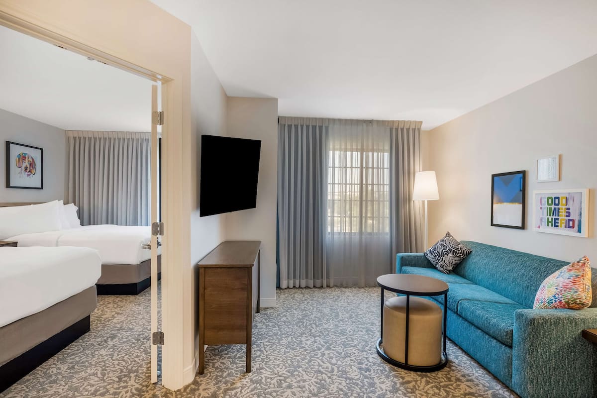 Habitación de hotel con dos camas, un sofá, un televisor, una lámpara y una alfombra estampada.
