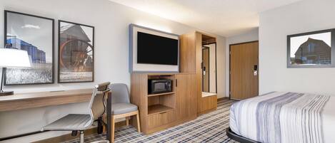 A hotel room with a bed, a desk with a computer, a chair, a TV, and a microwave.