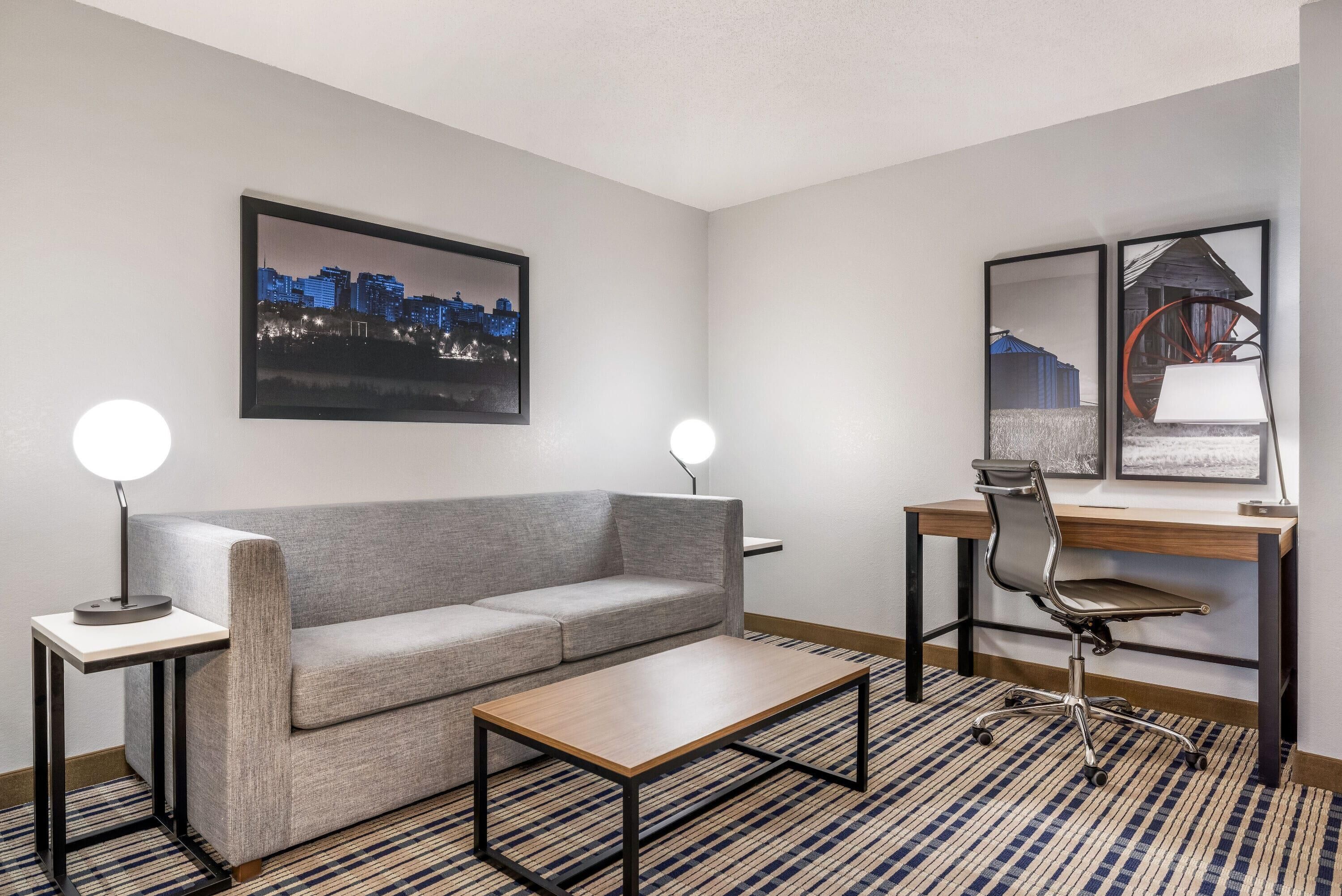 Una habitació d'hotel moderna amb un sofà gris, una taula baixa de fusta, un escriptori amb una cadira i dues làmpades penjades a la paret.