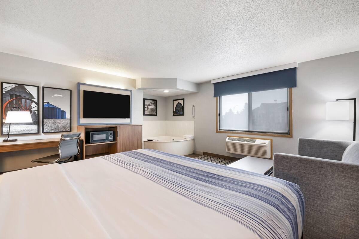 A modern hotel room with a large bed, a desk with a computer, a flat-screen TV, a bathtub, and a large window with a city view.