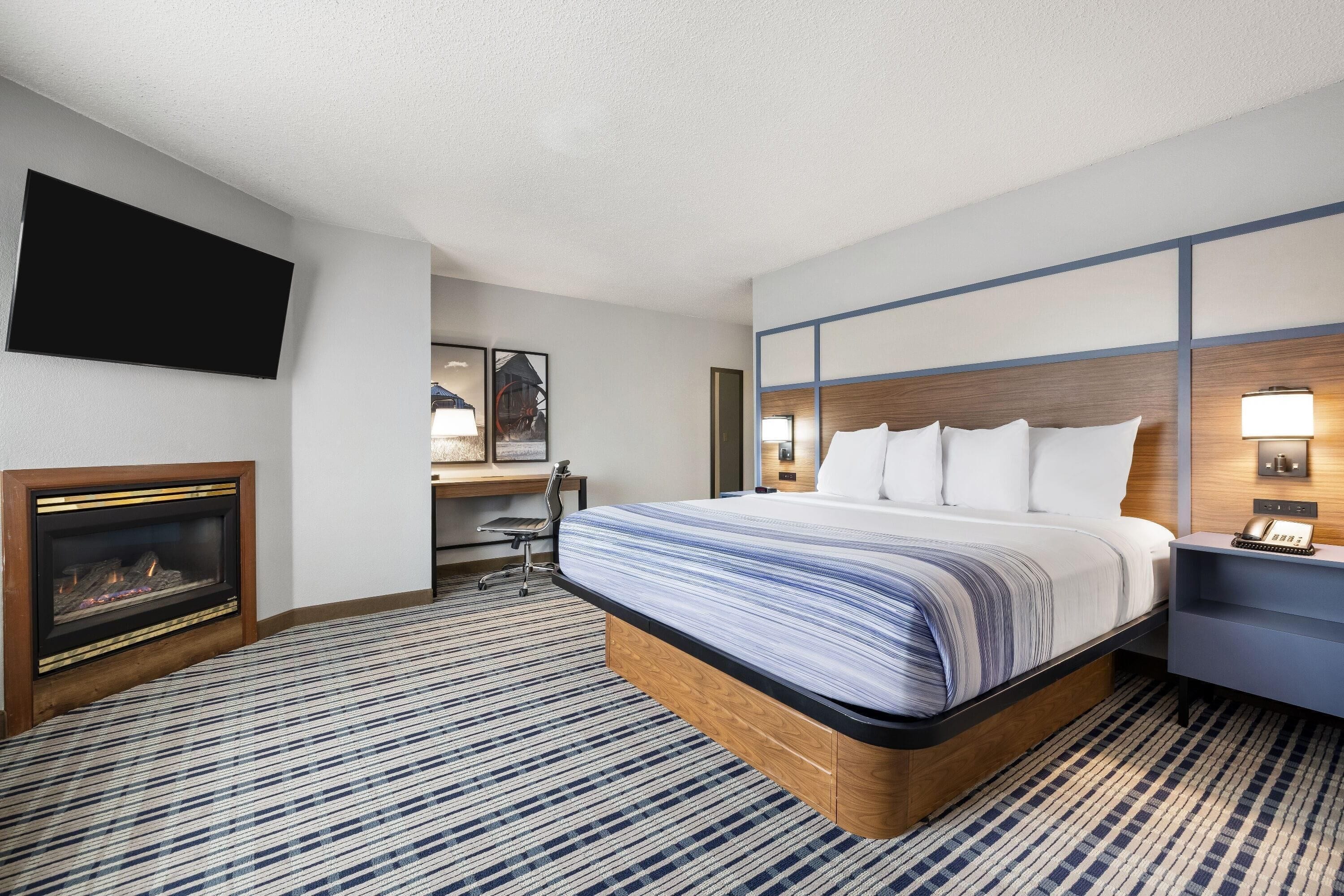 A hotel room with a large bed, a desk, a flat-screen TV, and a fireplace.