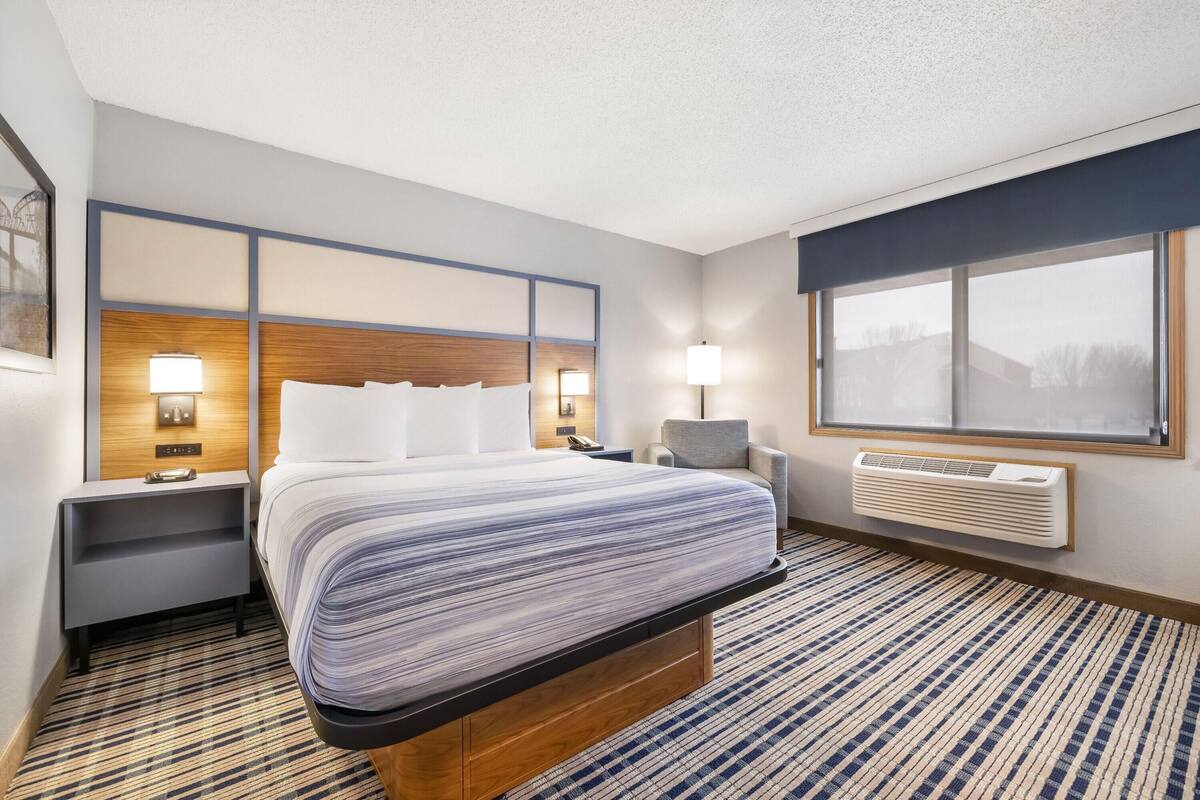 A hotel room with a large bed, bedside tables, a window, and a wall-mounted air conditioner.