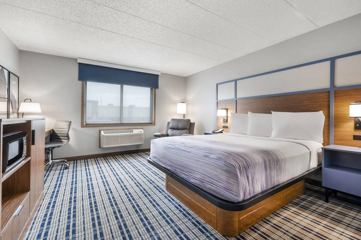 A hotel room with a large bed, a desk with a computer, a chair, a TV, and a window with blinds.