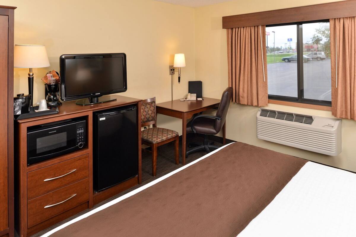 A hotel room with a bed, a desk with a chair, a television, a mini-fridge, a microwave, a lamp, and an air conditioning unit.