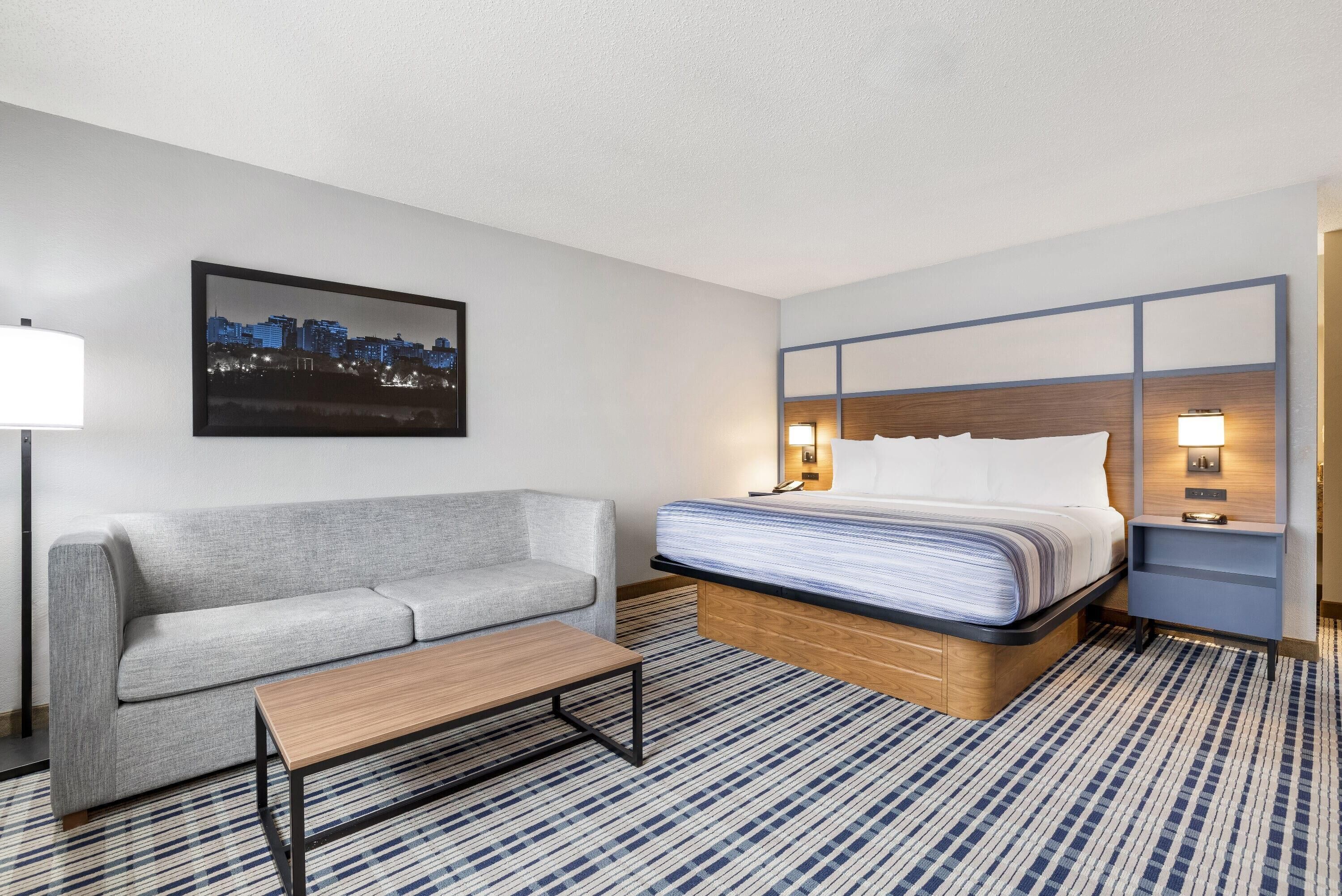 Habitación de hotel con cama, sofá, mesa de centro, un cuadro de paisaje urbano y alfombra estampada.