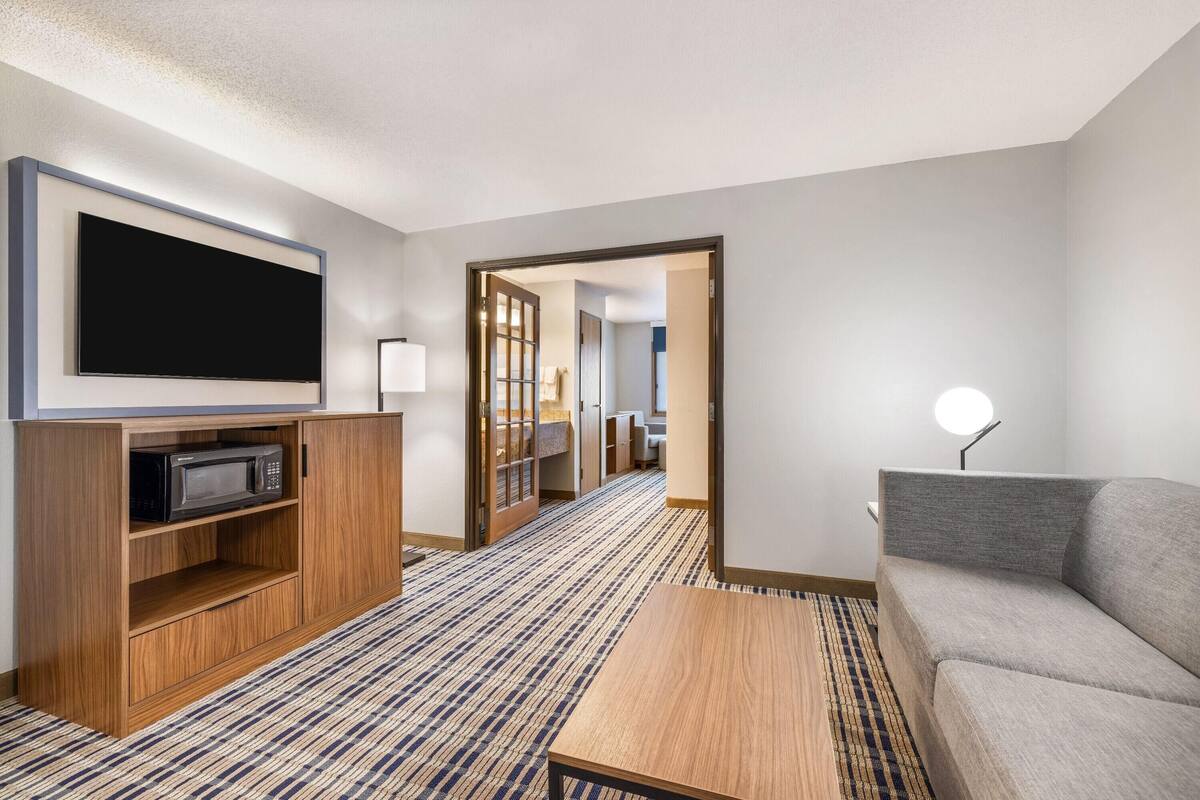 A modern hotel room with a flat-screen TV, a microwave, a sofa, a coffee table, and a hallway leading to a bathroom.