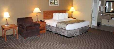 A hotel room with a bed, a chair, two bedside tables with lamps, a small table, a mirror, and a bathroom with a sink and toilet.