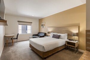 A hotel room with a large bed, a desk with a chair, a small table, and a window with blinds.