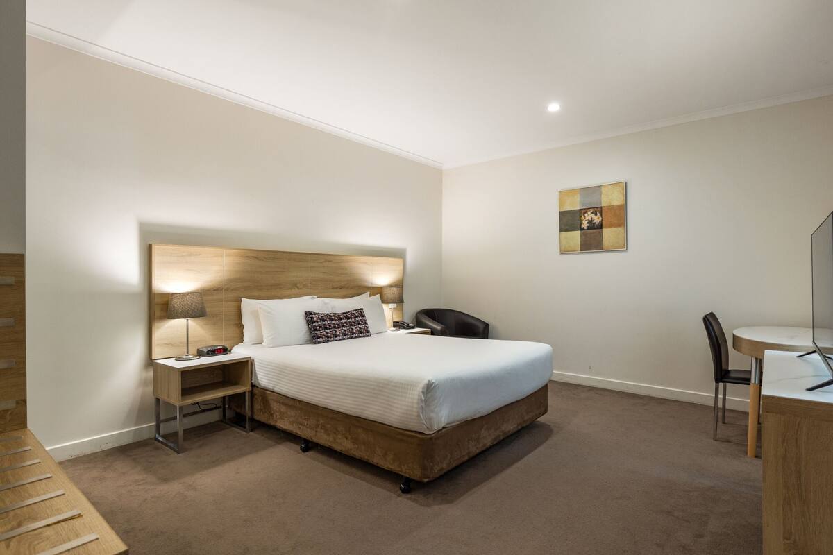 Quarto de hotel com cabeceira de madeira, cama com roupa de cama branca, mesa de cabeceira com abajur, escrivaninha com cadeira e uma pequena obra de arte na parede.