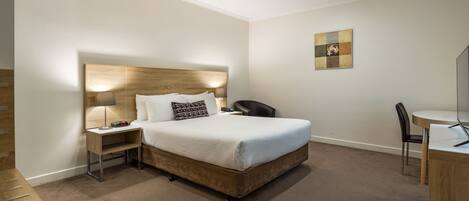 A hotel room with a wooden headboard, a bed with white linens, a bedside table with a lamp, a desk with a chair, and a small artwork on the wall.