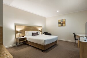 A hotel room with a wooden headboard, a bed with white linens, a bedside table with a lamp, a desk with a chair, and a small artwork on the wall.