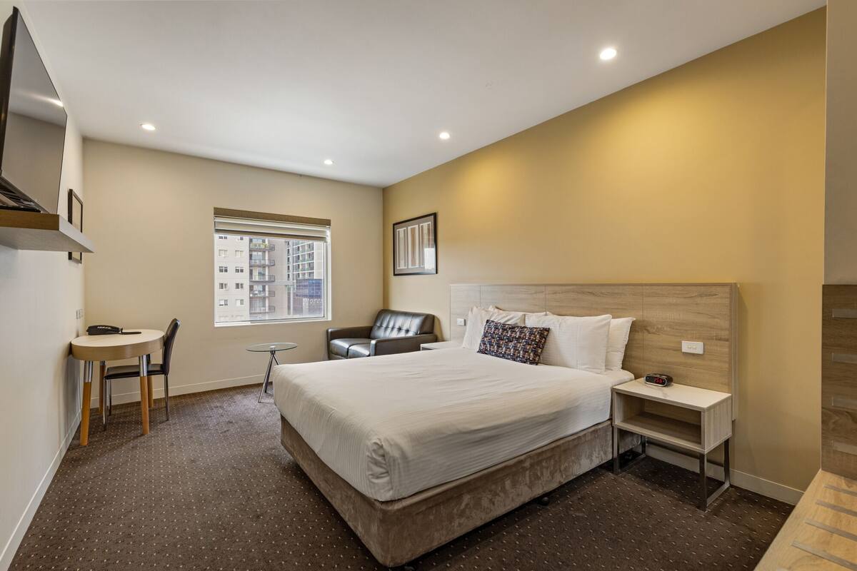 Quarto de hotel com uma cama grande, uma escrivaninha, uma cadeira, uma mesa pequena e uma janela com vista para edifícios.