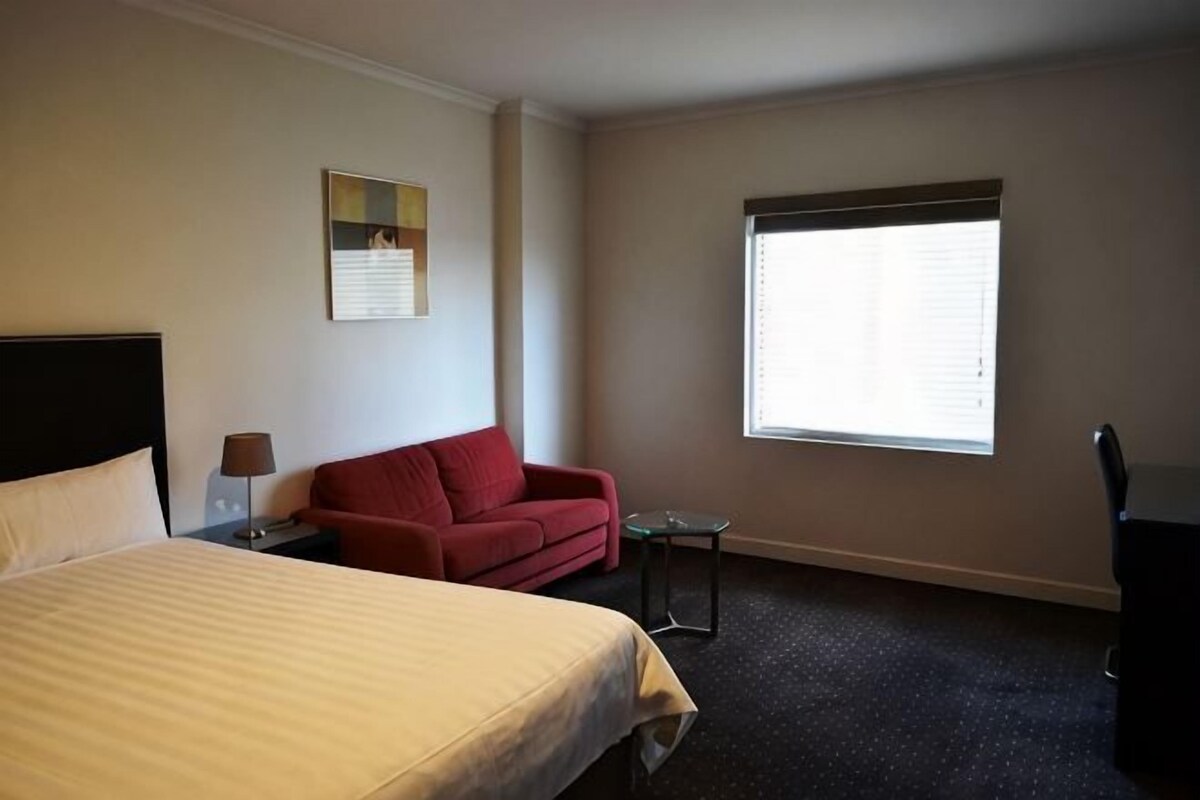 Quarto de hotel com cama, um sofá vermelho, uma mesa pequena, luminária, cadeira e janela com persianas.