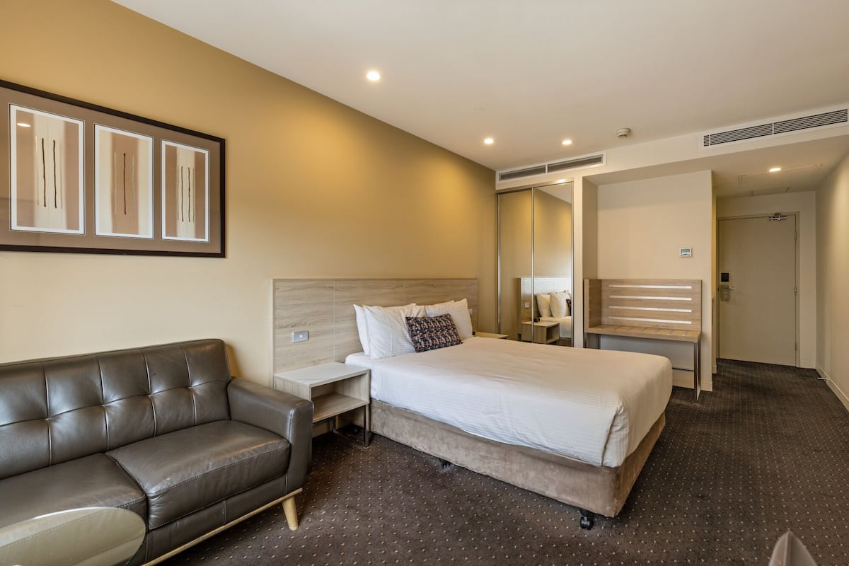 Quarto de hotel com uma cama, um sofá de couro, uma mesa de cabeceira, um espelho e uma porta.