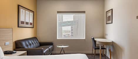 Habitación de hotel con cama, sofá de cuero, una mesita redonda y una silla. Se ve una ventana con vista a un puente y edificios.