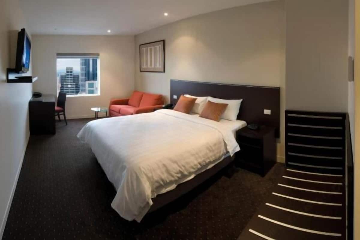 Quarto de hotel com cama, televisão, cadeira, mesa pequena e janela com vista para a cidade.