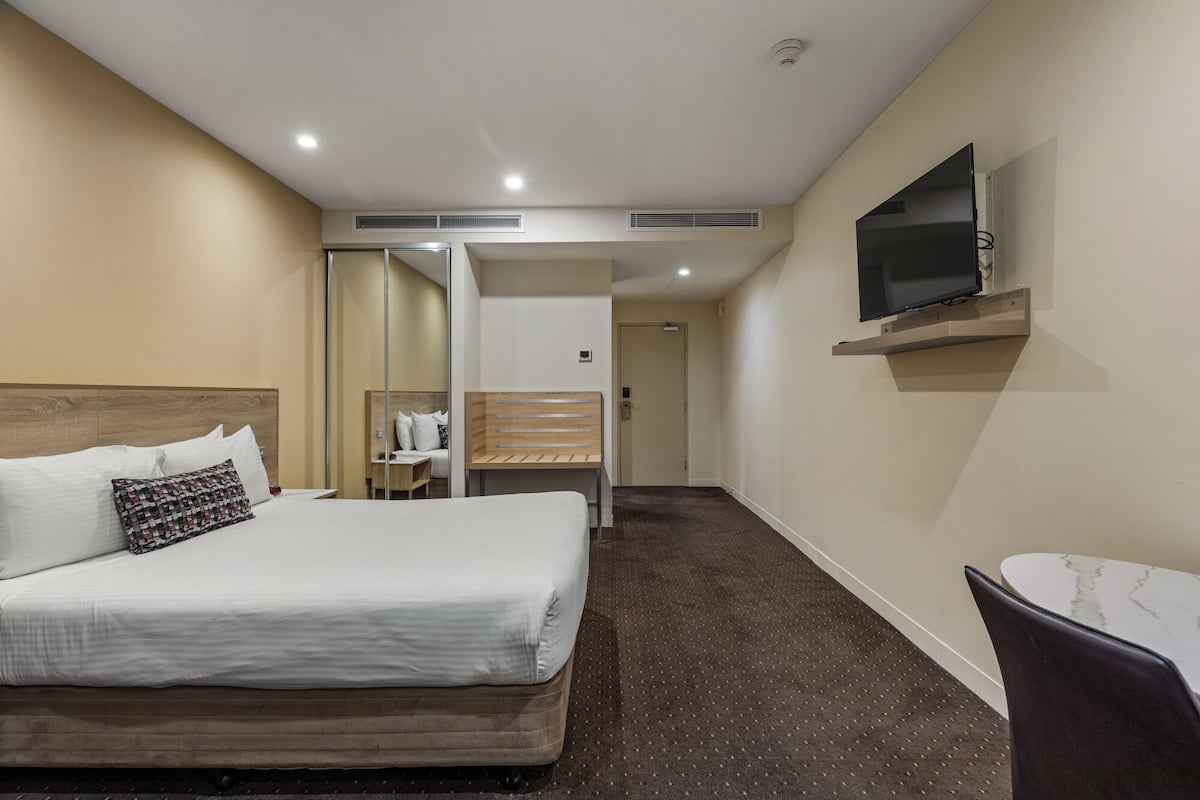 Quarto de hotel com uma cama, televisão na parede, uma mesinha e uma cadeira.