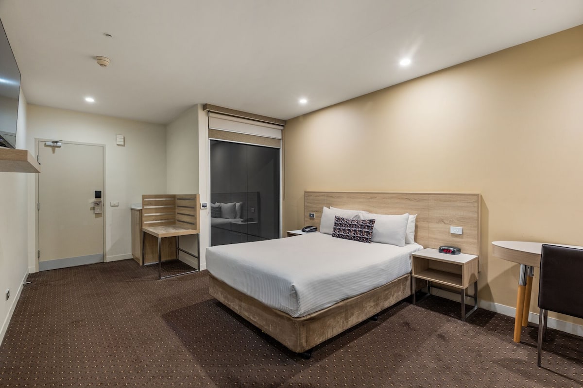 Quarto de hotel com uma cama grande, uma escrivaninha, uma cadeira, um guarda-roupa e uma mesinha.